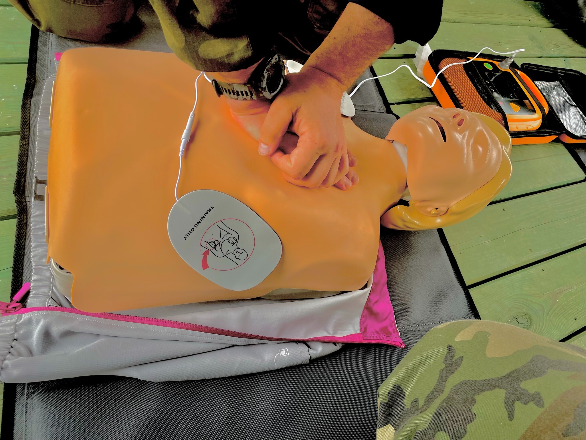 Instruktor wykonuje resuscytację krążeniowo-oddechową na manekinie z podłączonym defibrylatorem, widoczny zegarek na nadgarstku instruktora.