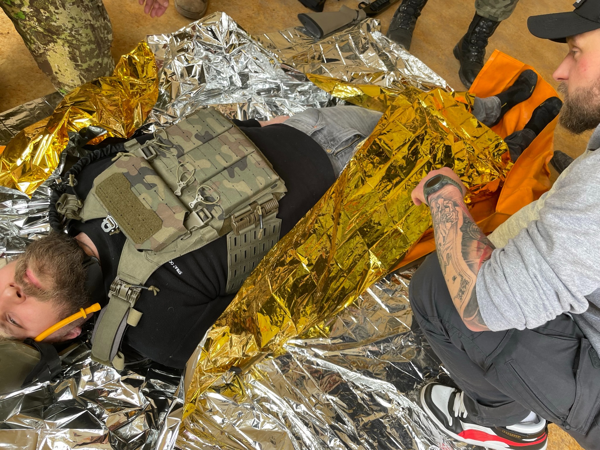 Osoba w kamizelce taktycznej leży na kocu termicznym, z rurką ustno-gardłową w ustach, instruktor z tatuażem na ramieniu udziela pomocy.