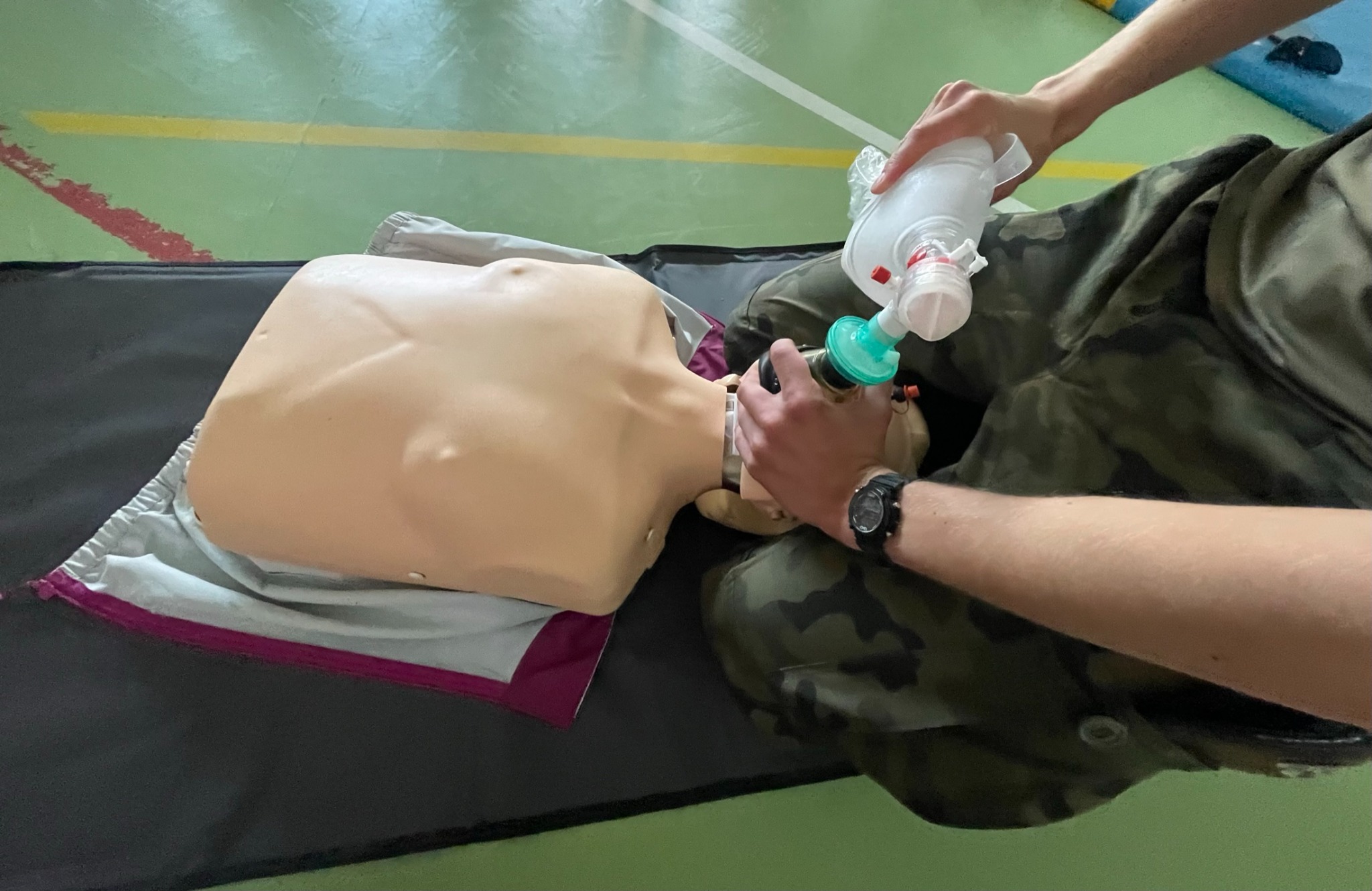 Instruktor w wojskowym kamuflażu demonstruje resuscytację na fantomie z użyciem worka samorozprężalnego na tle zielonej podłogi sali gimnastycznej.