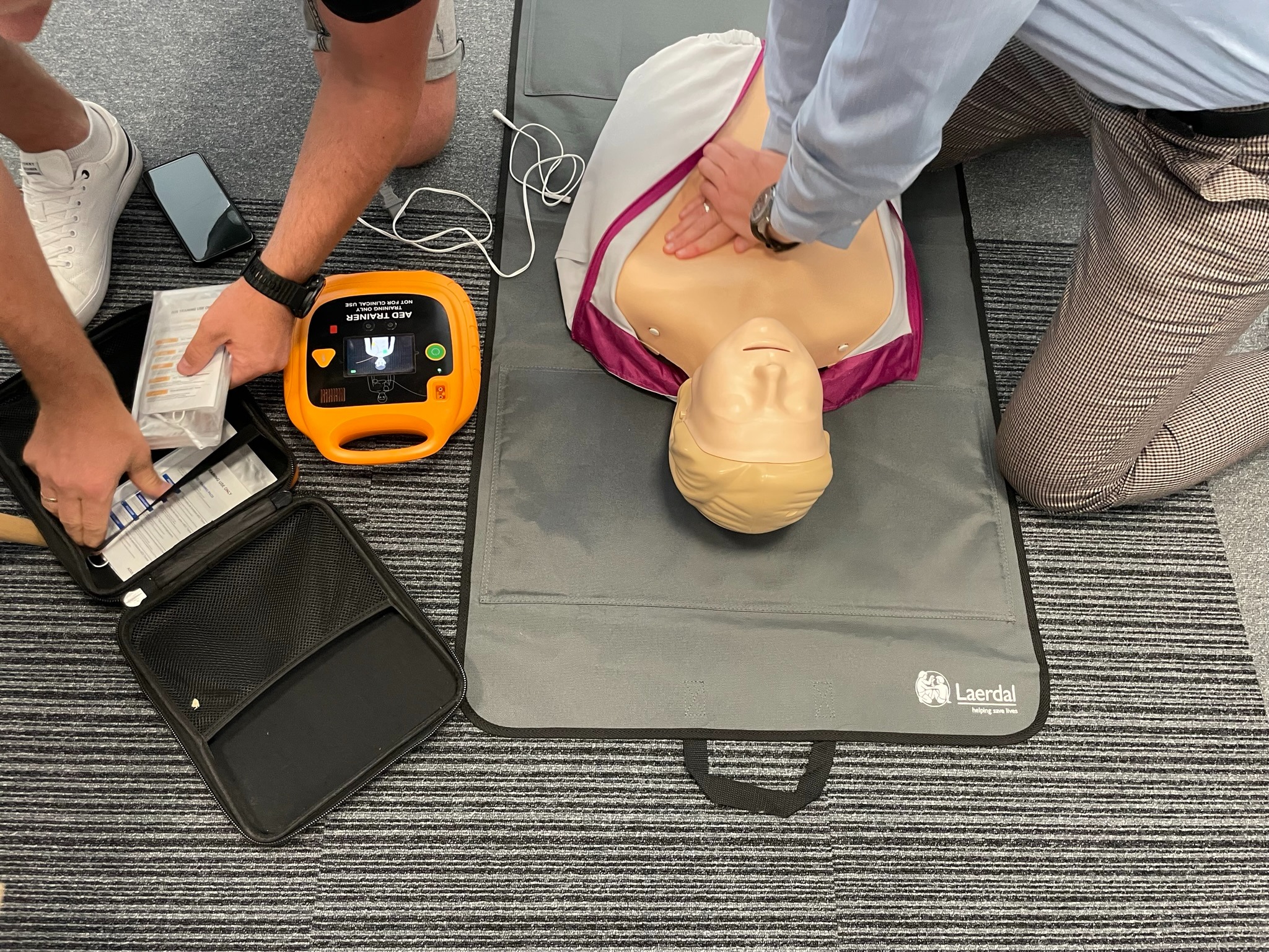 Instruktor demonstruje uciskanie klatki piersiowej na manekinie podczas szkolenia z pierwszej pomocy, obok leży defibrylator AED Trainer w pomarańczowej obudowie i otwarta torba z akcesoriami...