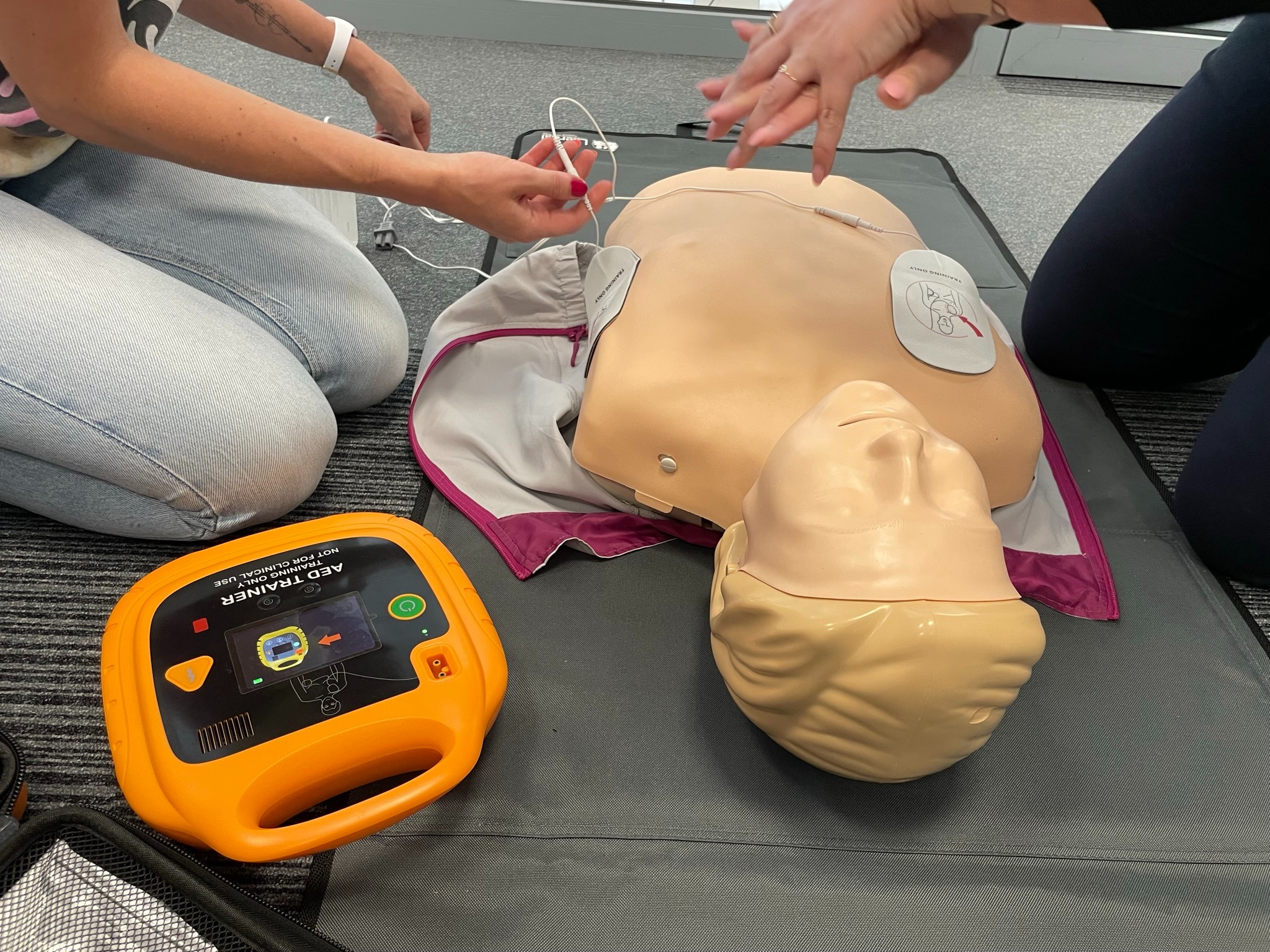 Instruktorzy podłączają elektrody do manekina podczas symulacji użycia defibrylatora AED Trainer; widoczny pomarańczowy defibrylator na szarym podłożu.