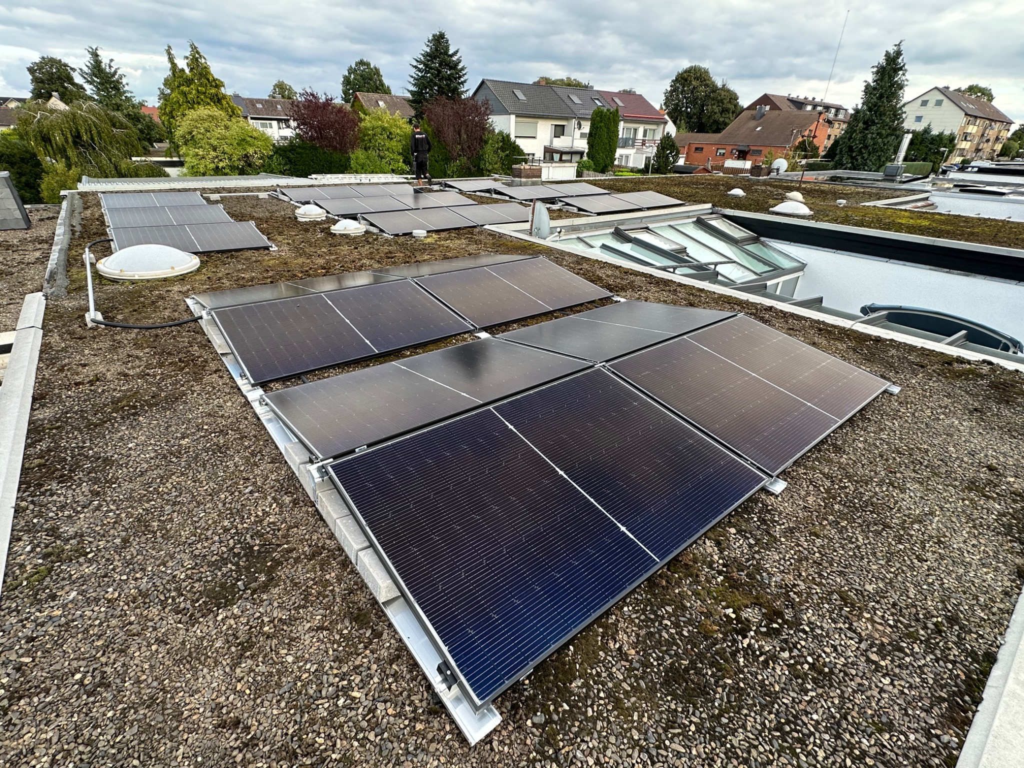 Instalacja paneli fotowoltaicznych na płaskim dachu pokrytym żwirem i mchem, z widocznym pracownikiem w oddali i zabudową osiedlową w tle pod pochmurnym niebem.