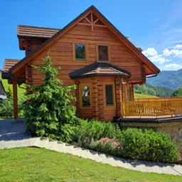 Dwukondygnacyjny dom z bali z drewnianym balkonem i kamienną podmurówką, otoczony zielenią, z widokiem na góry w tle.