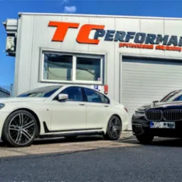 Dwa samochody BMW serii 7, biały i czarny, zaparkowane przed budynkiem TC Performance z pomarańczowym logo, sugerujące profesjonalny chip tuning.