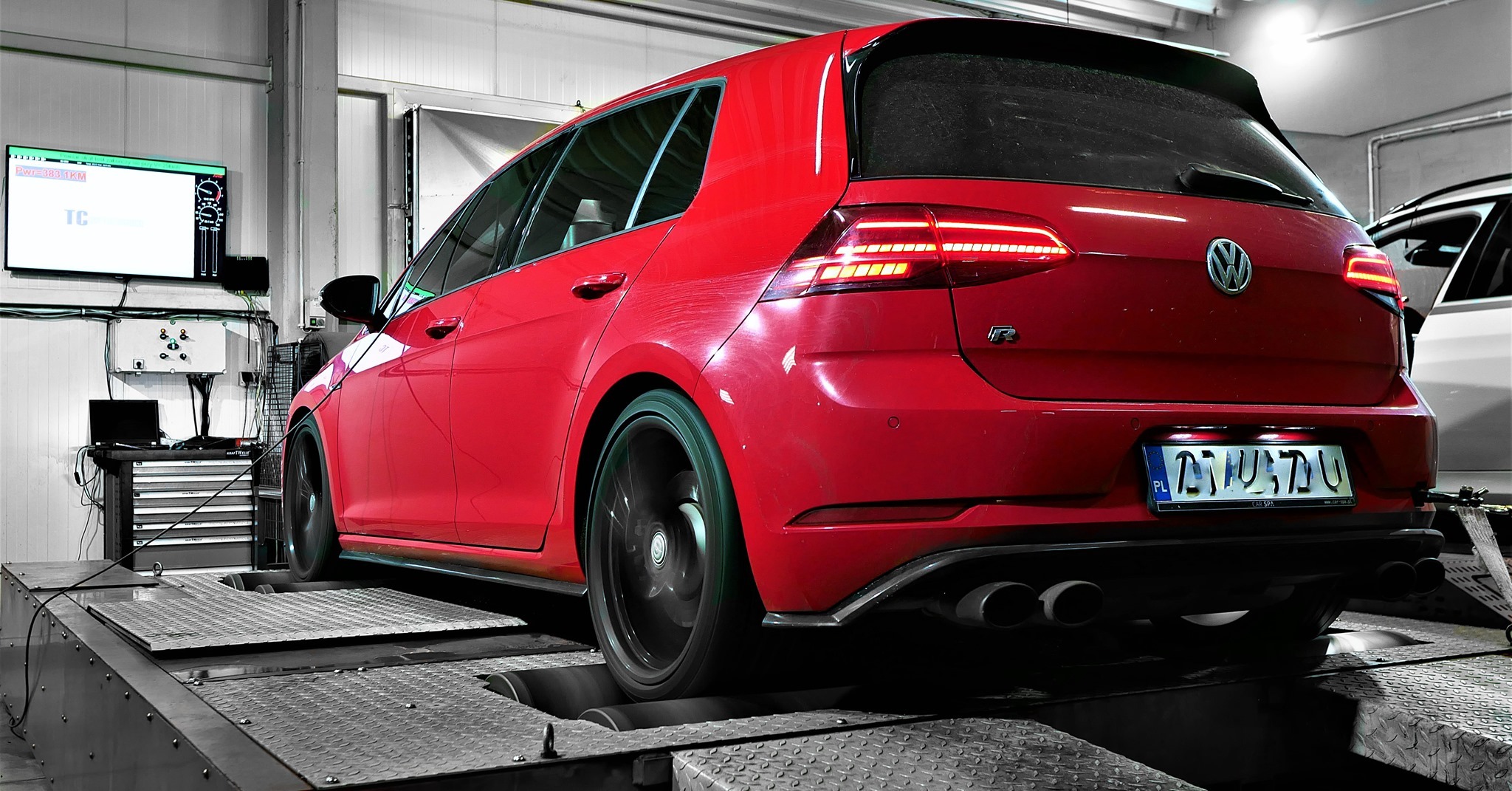 Czerwony samochód typu hatchback na hamowni podwoziowej, widoczne obracające się tylne koło, ekran z wynikami pomiarów mocy oraz stanowisko diagnostyczne w tle.