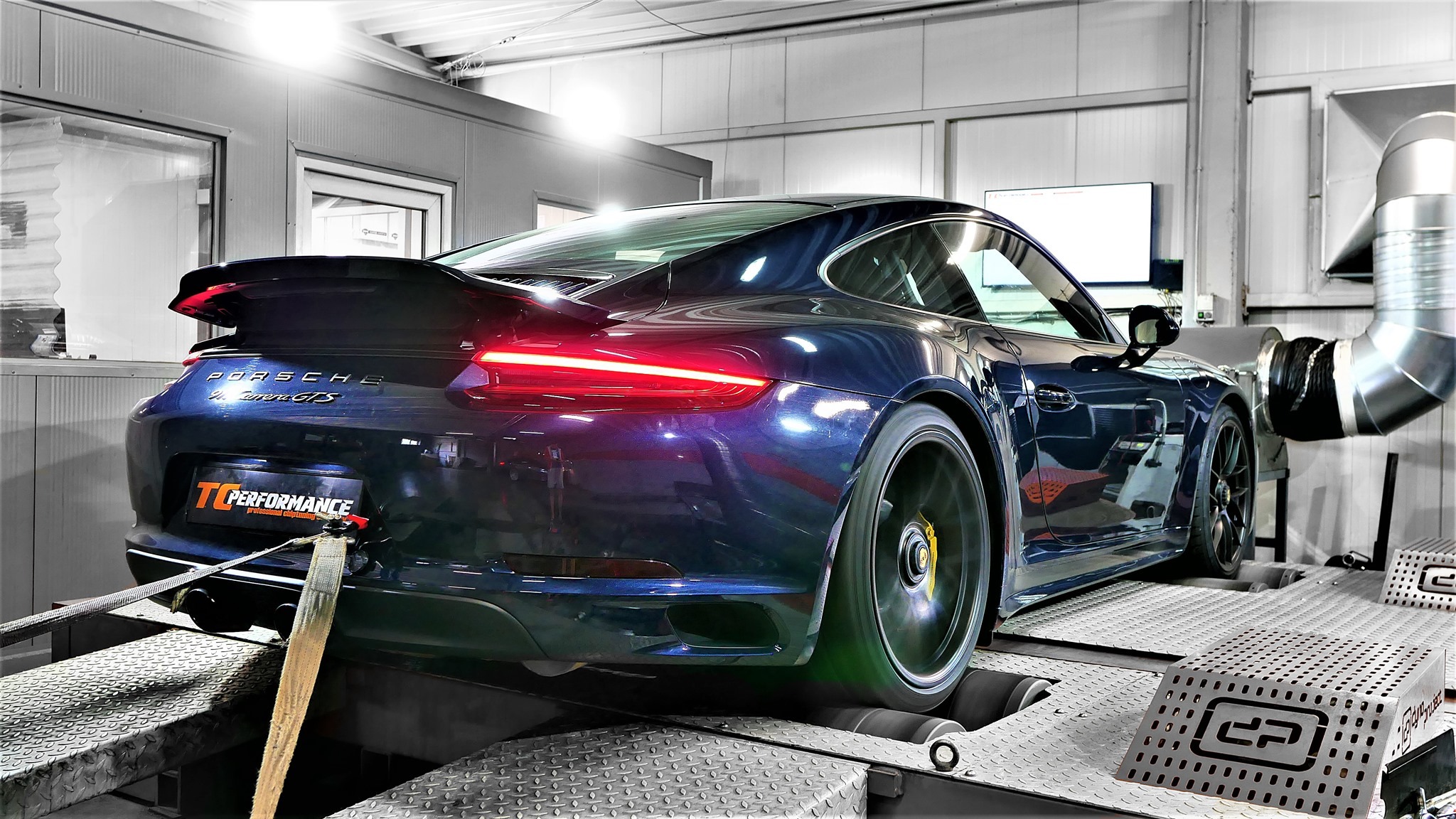 Granatowy Porsche Carrera GTS na hamowni podwoziowej, z widocznymi pasami mocującymi i obracającym się kołem, w tle białe ściany warsztatu.