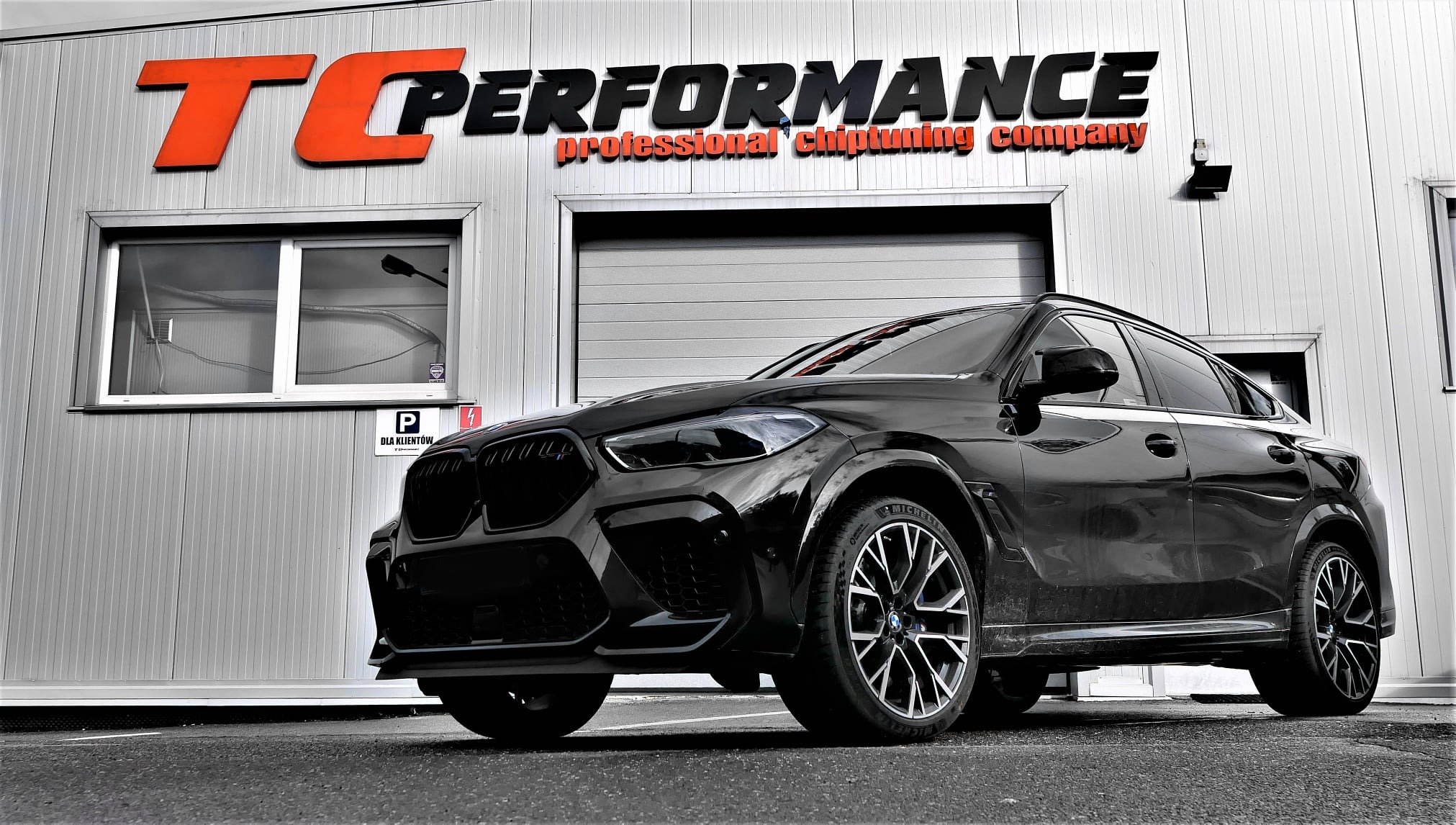 Czarny, sportowy SUV BMW stoi przed budynkiem firmy TC Performance, specjalizującej się w chiptuningu, z pomarańczowo-czarnym logo na fasadzie.
