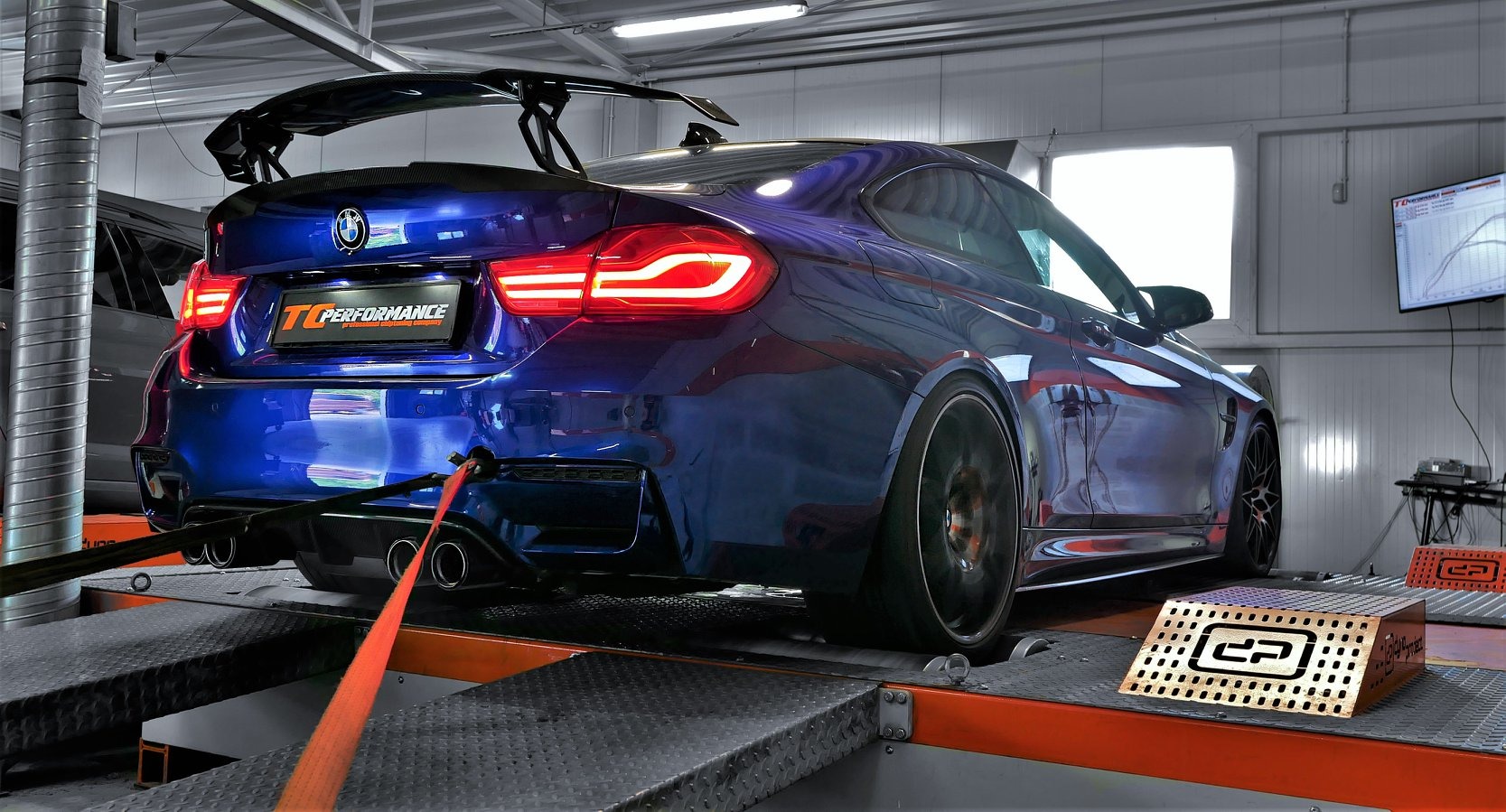Błękitne BMW M4 z karbonowym skrzydłem na hamowni podwoziowej, widoczne tylne światła, mocowania pasami i wykres na monitorze w tle.
