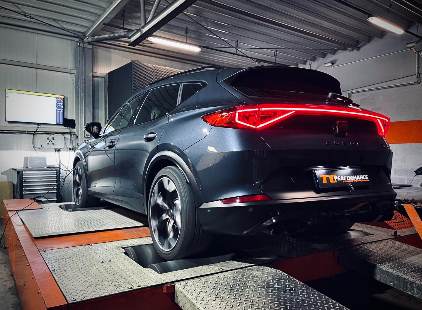 Szary samochód Cupra na hamowni podwoziowej; widoczne obracające się koła, pomarańczowe rolki hamowni, tylne światła LED, logo TC Performance i monitor z wykresami.