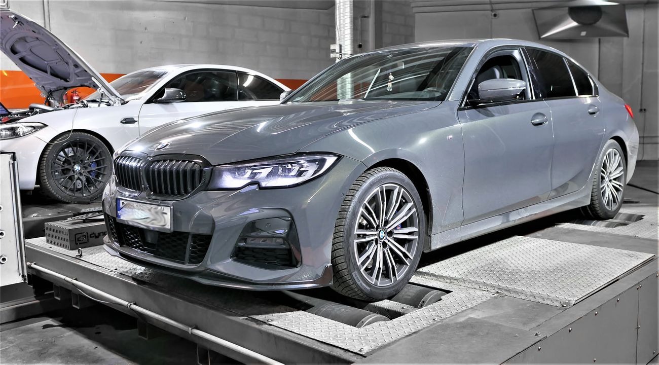 Szary sedan BMW na hamowni podwoziowej, z otwartą maską białego BMW w tle.