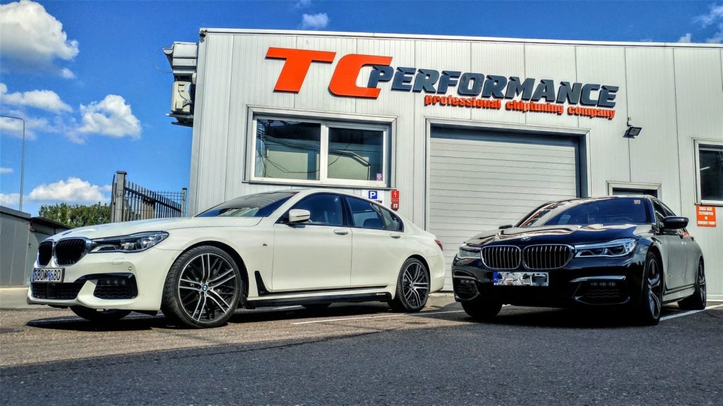 Dwa samochody BMW serii 7, biały i czarny, zaparkowane przed budynkiem TC Performance z pomarańczowym logo, sugerujące profesjonalny chip tuning.