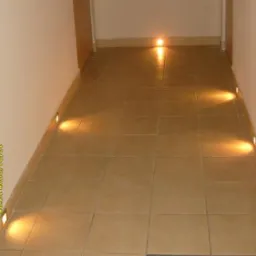 Korytarz z beżowymi płytkami podłogowymi i zamontowanymi wzdłuż ściany niskimi lampkami LED, dającymi ciepłe, punktowe światło.