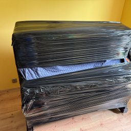 Przewoźnik Oleksandr Lisovytskyi - Pianino zabezpieczone czarną folią stretch przed transportem, widoczne drewniane nogi i fragment podłogi. Żółta ściana w tle.