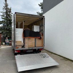 Przewoźnik Oleksandr Lisovytskyi - Bus transportowy z otwartą rampą załadowaną meblami, zabezpieczonymi folią i kocem. W tle siedząca osoba i drzewo iglaste.