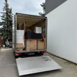 Bus transportowy z otwartą rampą załadowaną meblami, zabezpieczonymi folią i kocem. W tle siedząca osoba i drzewo iglaste.