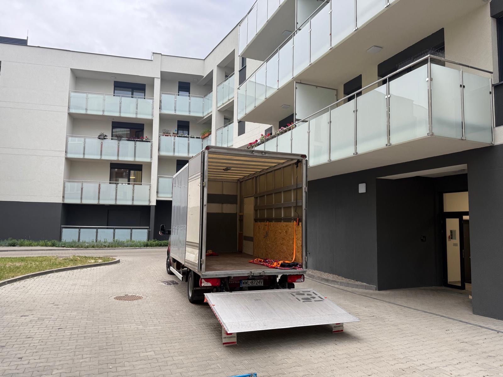 Otwarty bus do przeprowadzek z rampą zaparkowany przed nowoczesnym budynkiem mieszkalnym z balkonami i szklanymi balustradami. Widok z tyłu pojazdu.