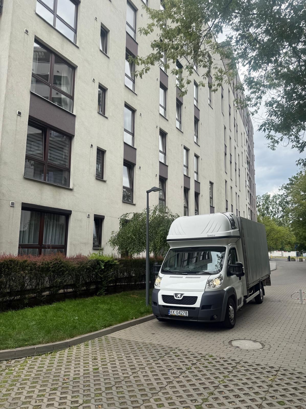 Biały bus z plandeką marki Peugeot zaparkowany przed nowoczesnym, wielopiętrowym budynkiem mieszkalnym z brązowymi ramami okiennymi. Kostka brukowa i zieleń.