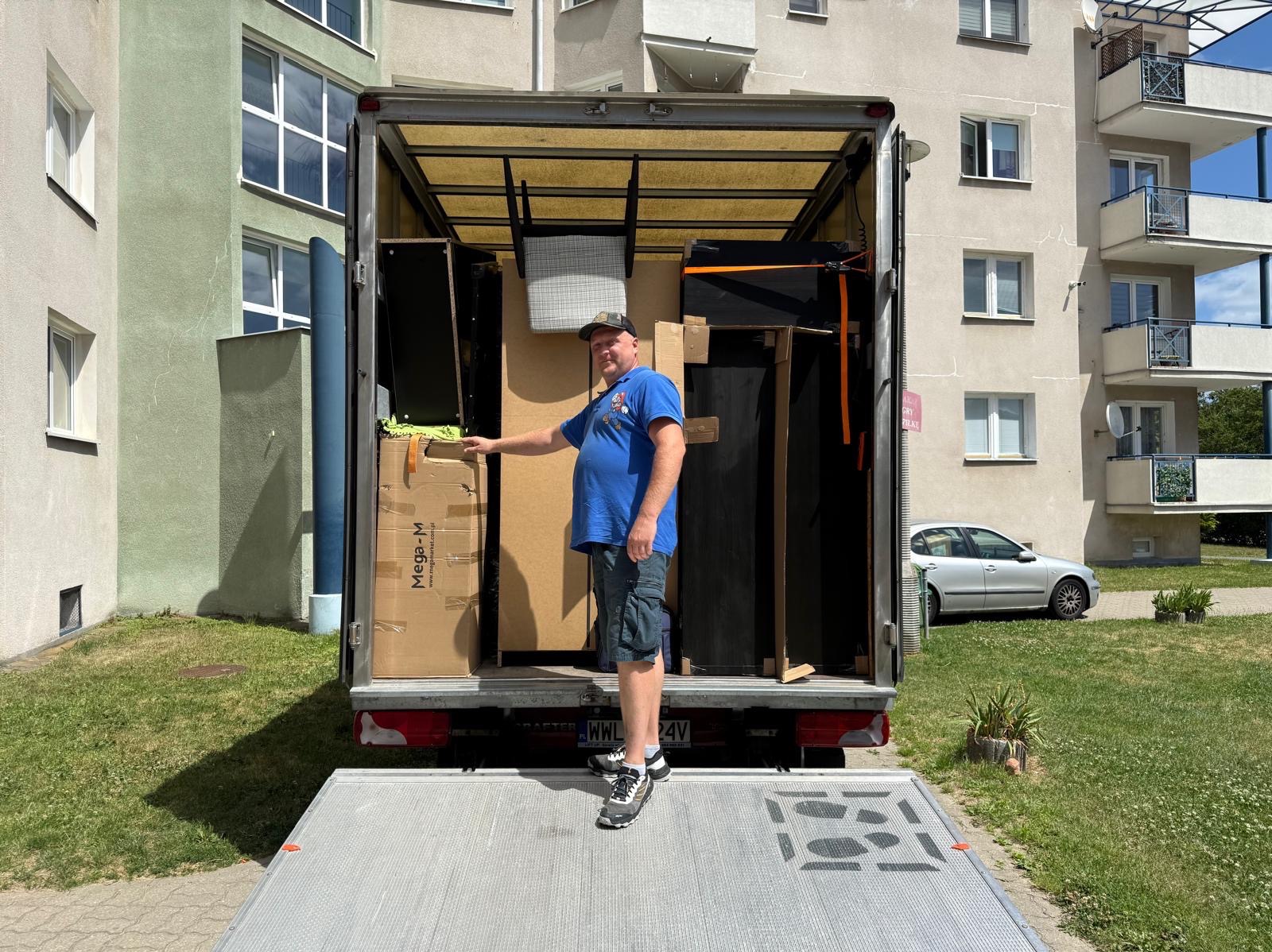 Mężczyzna przy załadowanym samochodzie dostawczym, gotowym do przeprowadzki, na tle bloku mieszkalnego i zielonego trawnika.