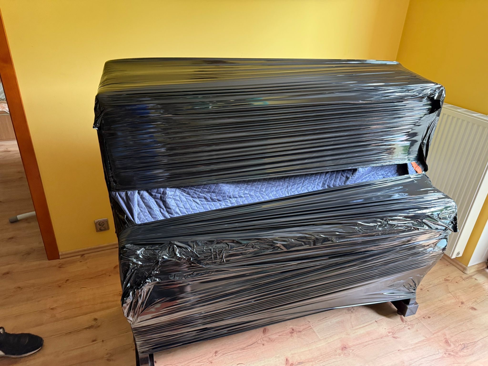 Pianino zabezpieczone czarną folią stretch przed transportem, widoczne drewniane nogi i fragment podłogi. Żółta ściana w tle.