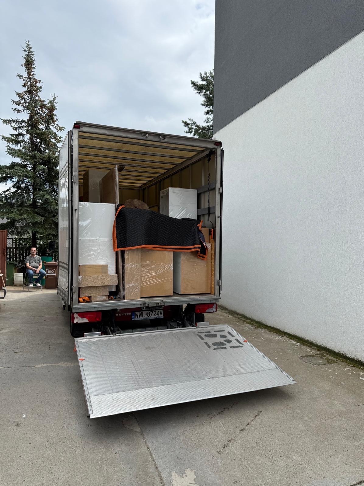 Bus transportowy z otwartą rampą załadowaną meblami, zabezpieczonymi folią i kocem. W tle siedząca osoba i drzewo iglaste.