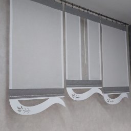 Firma Ewa - Trzy szare rolety rzymskie z białymi, dekoracyjnymi, wycinanymi laserowo falbanami z motywem liści, zawieszone na metalowym karniszu z klipsami, na tle kremowej ściany.