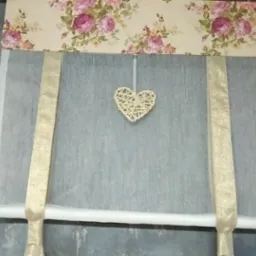 Delikatna roleta rzymska z kremową tkaniną w kwiaty i ozdobnym, plecionym sercem zawieszonym na środku.