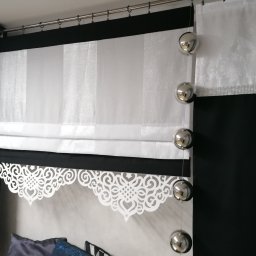 Firma Ewa - Elegancka aranżacja okienna: Biała roleta rzymska z dekoracyjnym, ażurowym wykończeniem, zestawiona z czarnymi zasłonami zdobionymi srebrnymi pasami i białym lambrekinem z motywem kwiatowym...