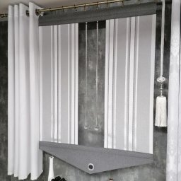 Firma Ewa - Nowoczesna roleta rzymska w paski, z szarym wykończeniem, zawieszona obok białych zasłon na metalowym karniszu, uzupełniona dekoracyjnymi elementami na tle szarej ściany.