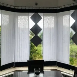 Nowoczesne, białe, pionowe żaluzje z czarnymi, geometrycznymi dekoracjami w kształcie rombów, widok na zielony ogród za oknem.