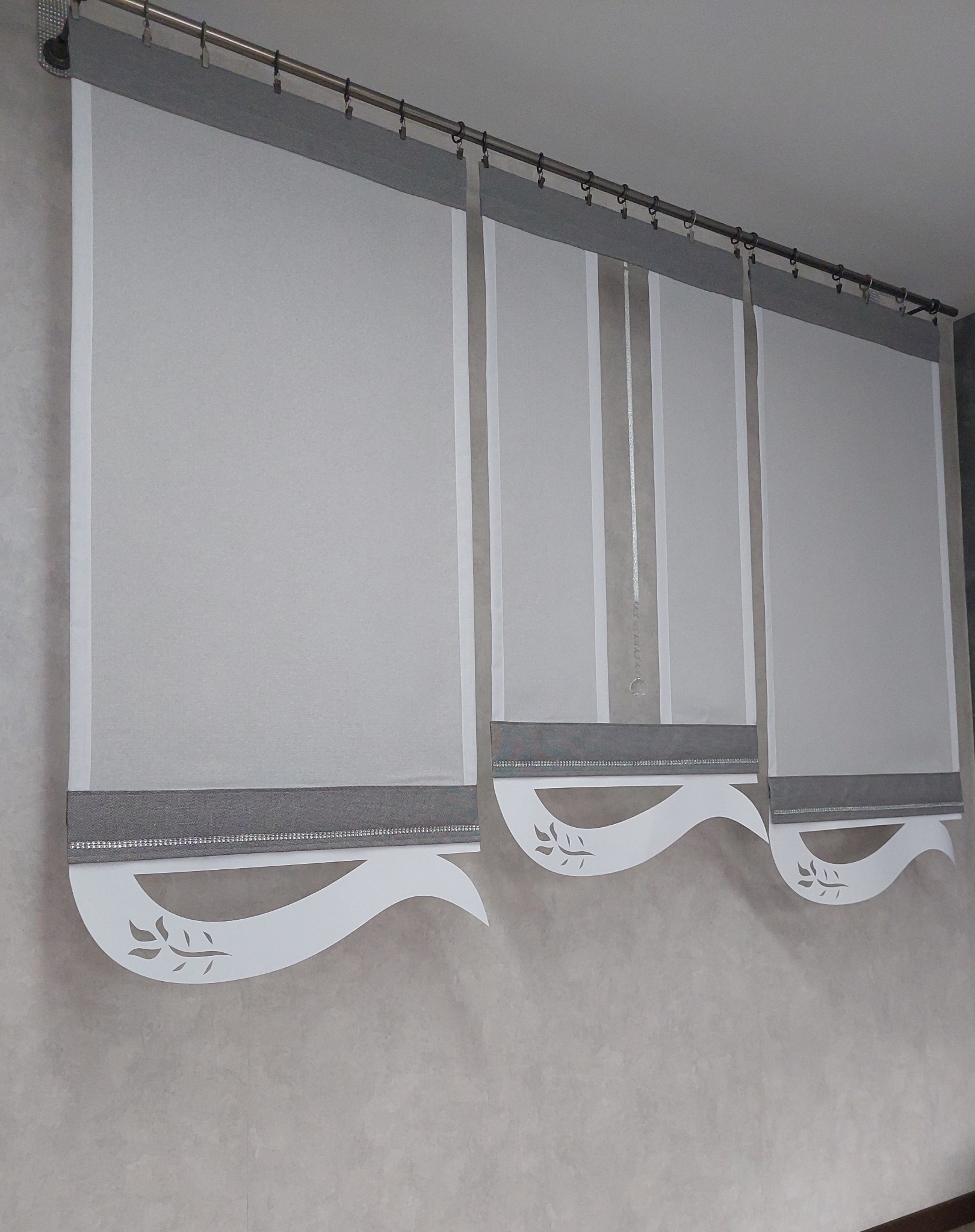 Trzy szare rolety rzymskie z białymi, dekoracyjnymi, wycinanymi laserowo falbanami z motywem liści, zawieszone na metalowym karniszu z klipsami, na tle kremowej ściany.