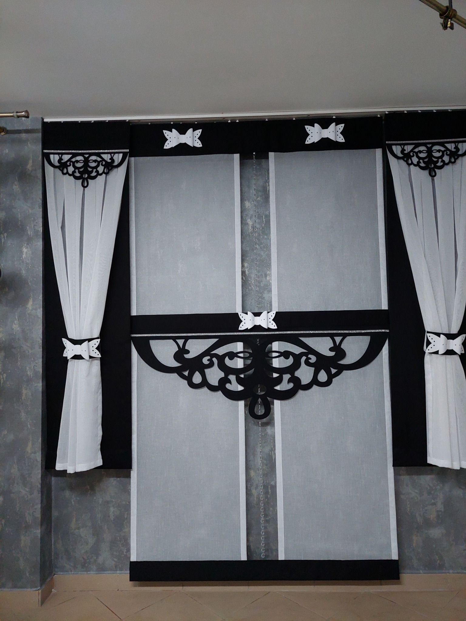 Eleganckie, biało-czarne zasłony z dekoracyjnymi kokardkami i ażurowymi elementami, zawieszone na karniszu w pomieszczeniu z szarą ścianą.