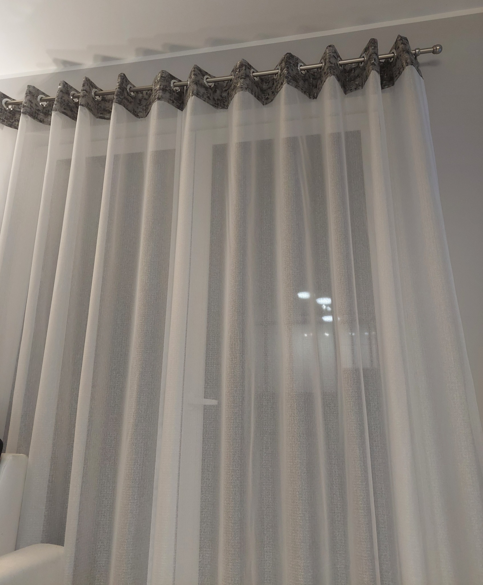 Delikatne, białe zasłony woalowe z szarymi akcentami w górnej części, zawieszone na metalowym karniszu z przelotkami, subtelnie przepuszczają światło.