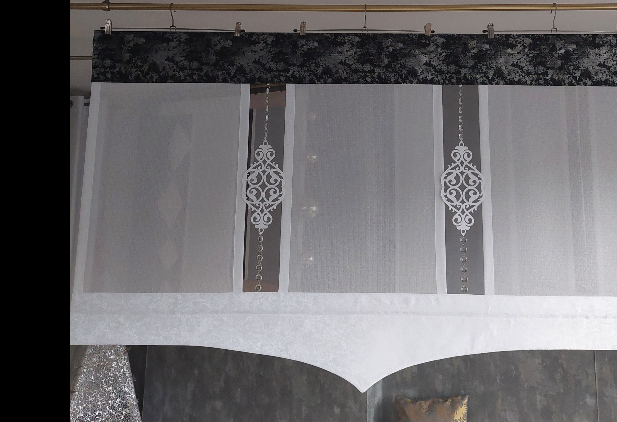 Biała, zwiewna zasłona z dekoracyjnymi, ażurowymi aplikacjami i srebrnymi koralikami, zawieszona na karniszu z czarnym, wzorzystym pasem materiału.