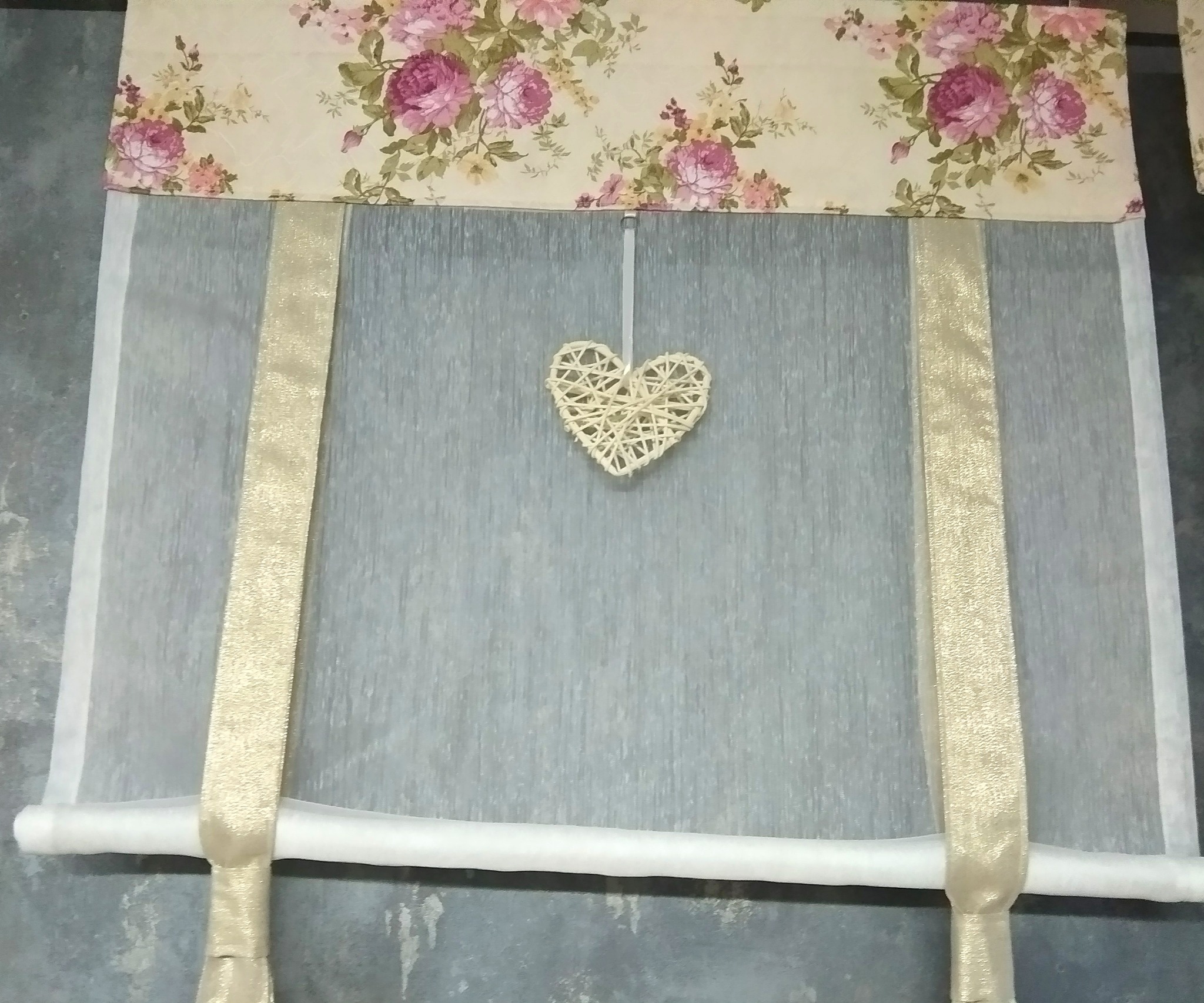 Delikatna roleta rzymska z kremową tkaniną w kwiaty i ozdobnym, plecionym sercem zawieszonym na środku.