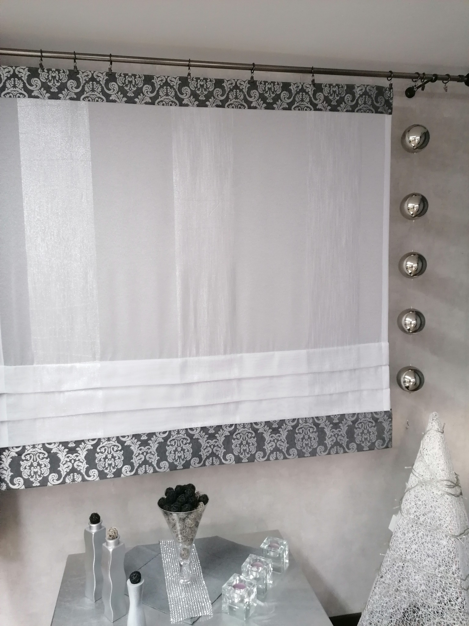 Elegancka roleta rzymska z białego, prześwitującego materiału, zdobiona szarym, ornamentowym wzorem na górze i dole, zawieszona na metalowym karniszu, z dekoracyjnymi srebrnymi kulami po prawej...