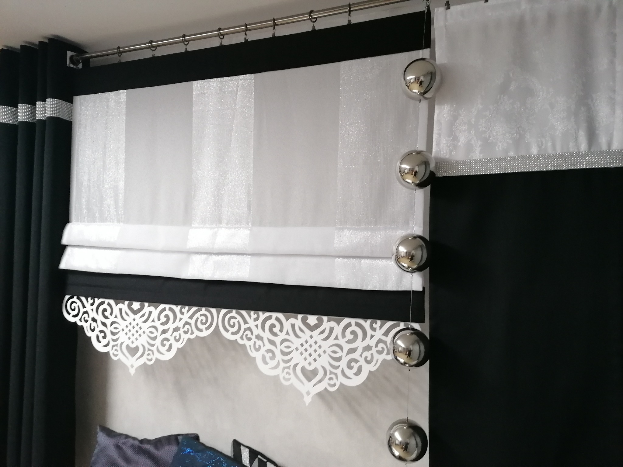 Elegancka aranżacja okienna: Biała roleta rzymska z dekoracyjnym, ażurowym wykończeniem, zestawiona z czarnymi zasłonami zdobionymi srebrnymi pasami i białym lambrekinem z motywem kwiatowym...
