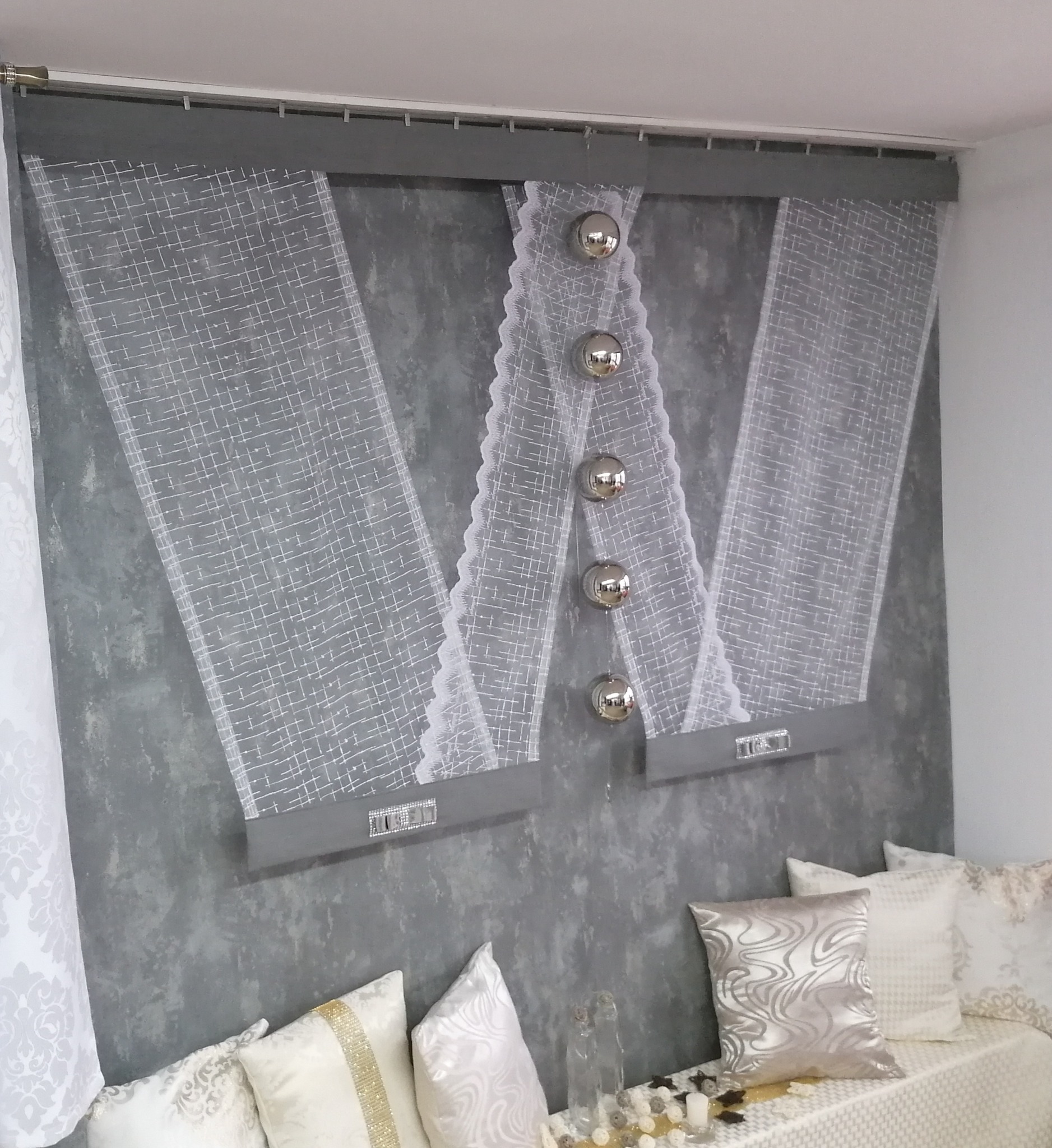 Nowoczesna aranżacja okienna z białych, wzorzystych zasłon panelowych z szarymi pasami i dekoracyjnymi srebrnymi kulami, dopełniona poduszkami w odcieniach bieli i srebra na tle szarej ściany.