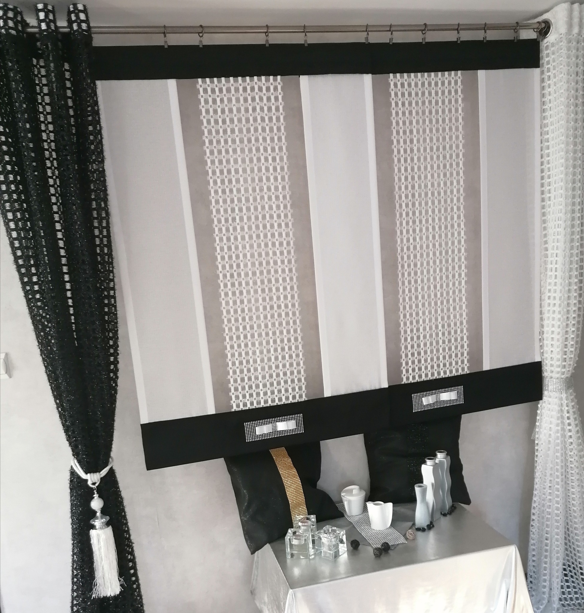 Dekoracja okienna z czarnych i białych zasłon panelowych z geometrycznym wzorem, ozdobionych kryształkami, z czarnymi poduszkami i srebrną dekoracją na stole.
