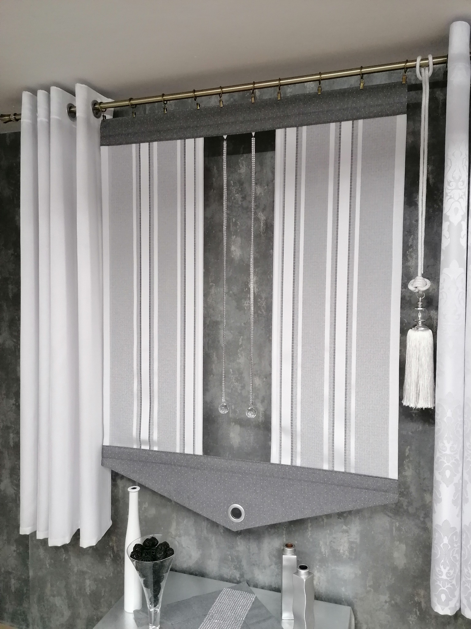 Nowoczesna roleta rzymska w paski, z szarym wykończeniem, zawieszona obok białych zasłon na metalowym karniszu, uzupełniona dekoracyjnymi elementami na tle szarej ściany.