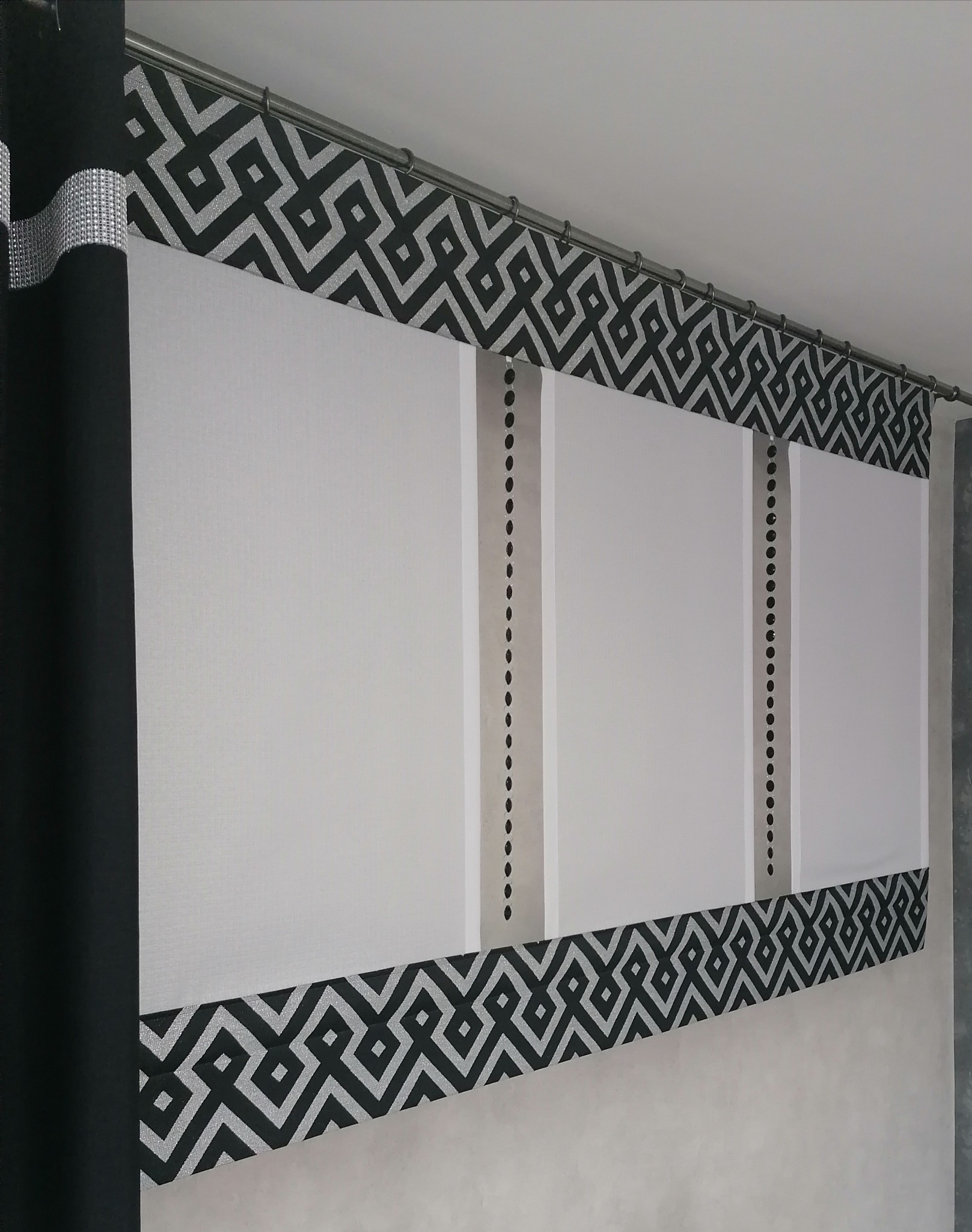 Roleta rzymska z geometrycznym wzorem w kolorze czarnym i srebrnym, widoczna z dołu, z dekoracyjnymi czarnymi zasłonami po lewej stronie.