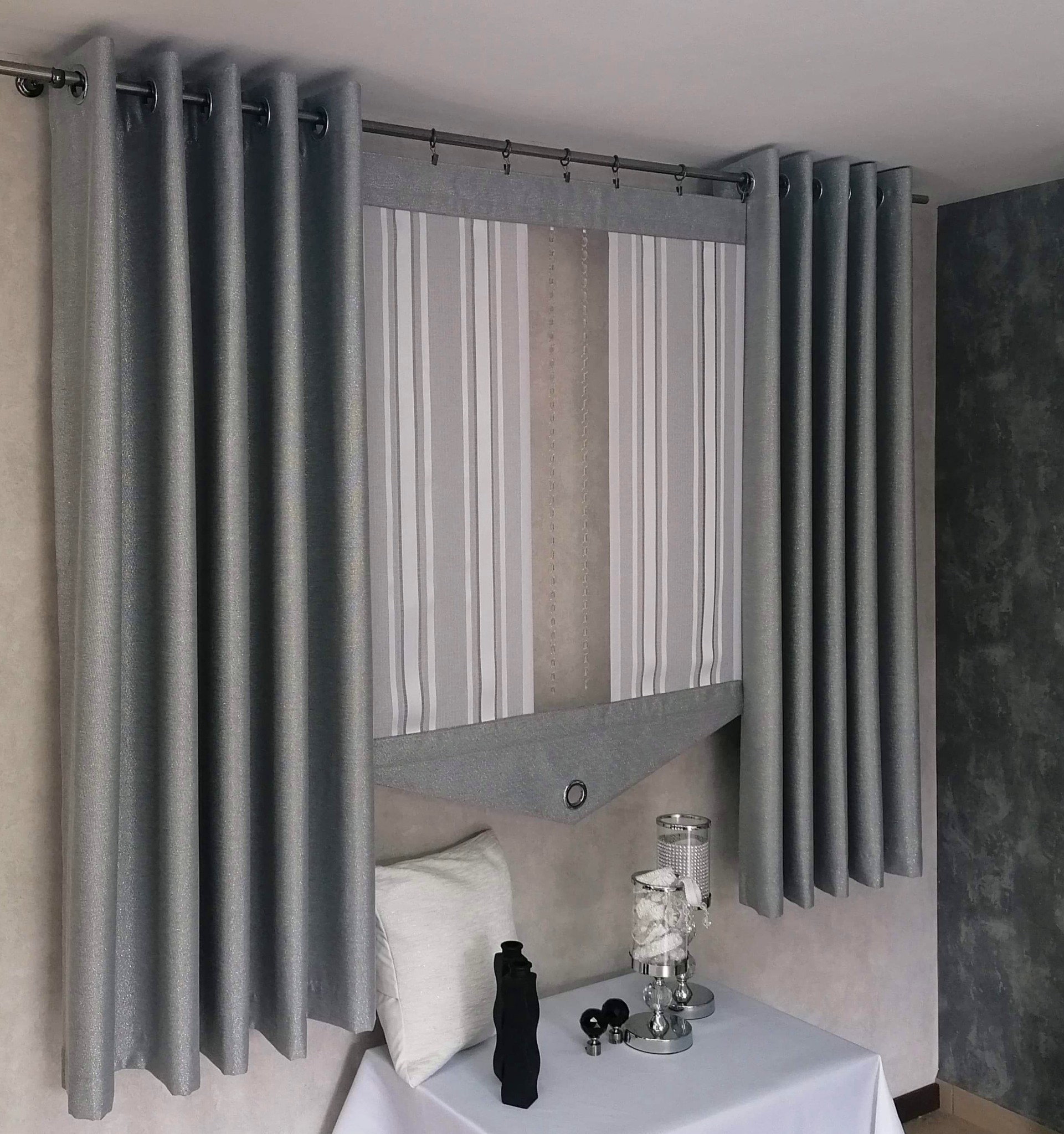 Szare, błyszczące zasłony panelowe z metalowymi przelotkami, po bokach zasłony z tego samego materiału, w tle roleta rzymska w biało-szare pasy, na stole dekoracje i poduszka.