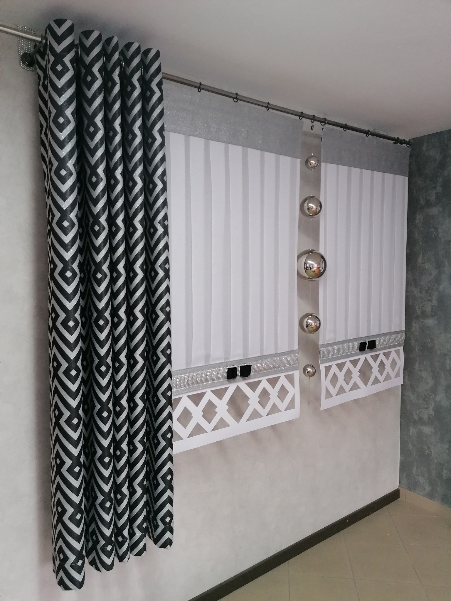 Czarno-białe zasłony w geometryczny wzór wiszące obok białych rolet rzymskich z dekoracyjnymi srebrnymi elementami i ozdobnymi kulami.