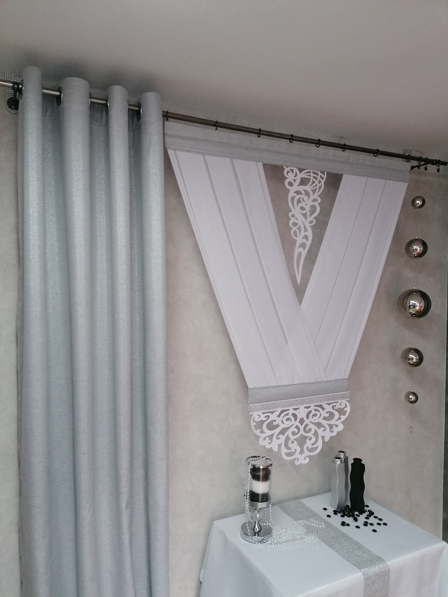 Elegancka aranżacja okienna: srebrne zasłony z metalicznym połyskiem i dekoracyjna, biała firana w kształcie litery V, wykończona ażurowym wzorem i błyszczącą taśmą, uzupełniona srebrnymi kulami...