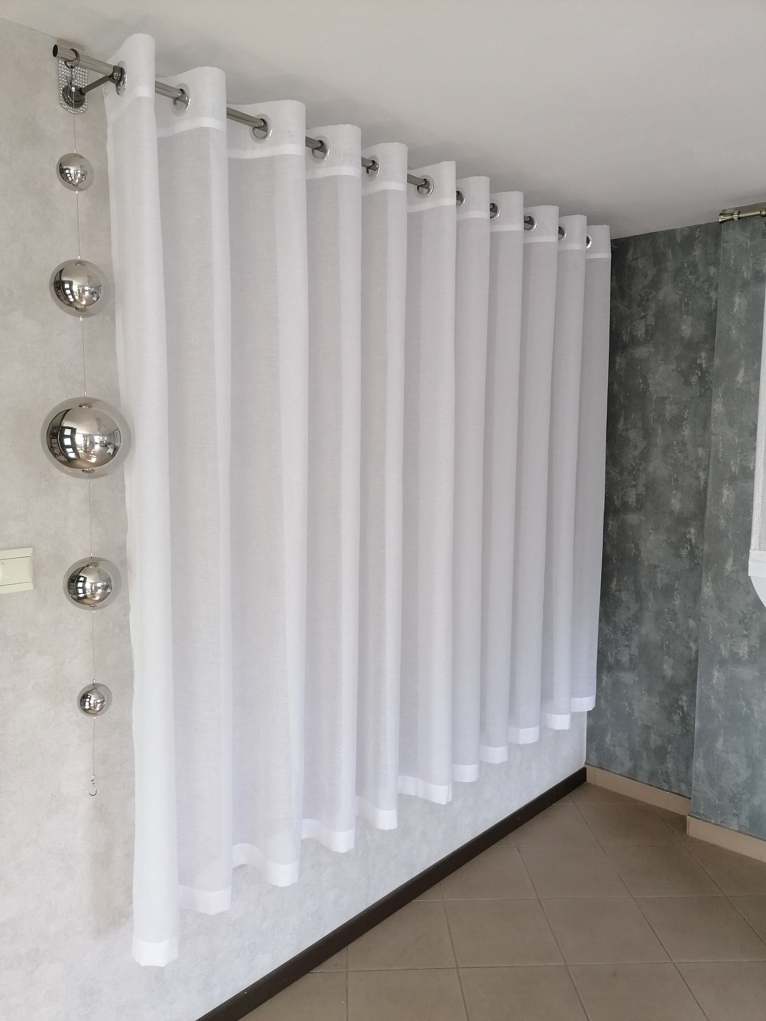 Długie, białe, prześwitujące zasłony z metalowymi kółkami zawieszone na karniszu, z dekoracją w postaci srebrnych kul.