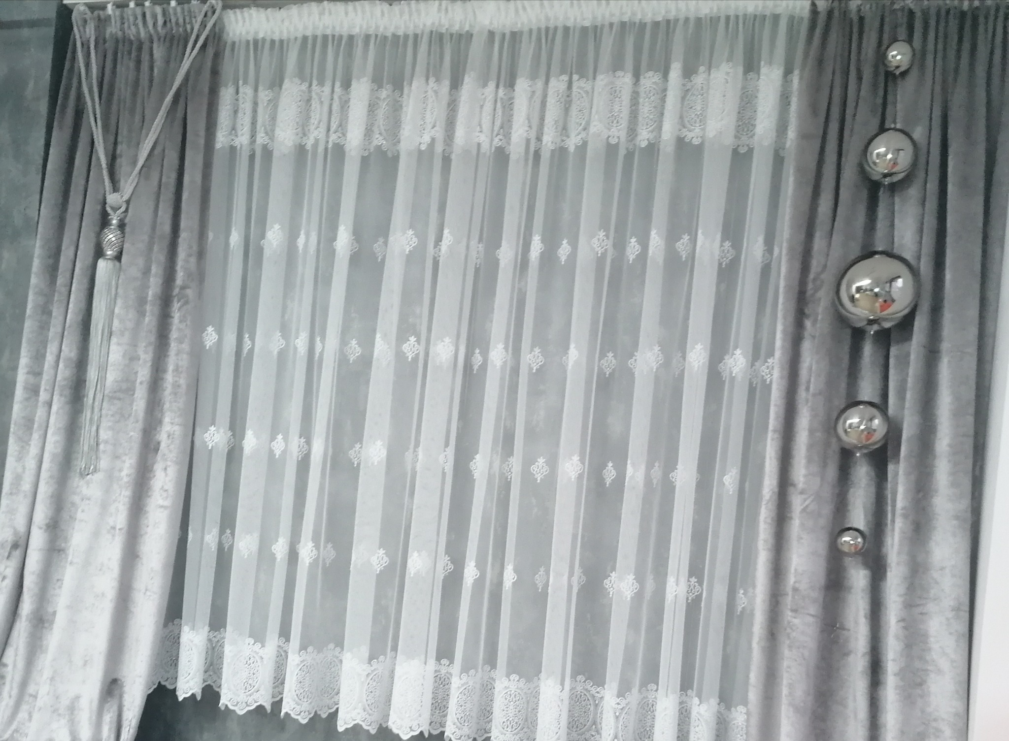 Szare, welurowe zasłony z dekoracyjnym sznurem i srebrnym chwostem, zestawione z delikatną, białą firanką z haftowanym wzorem i srebrnymi kulami dekoracyjnymi.