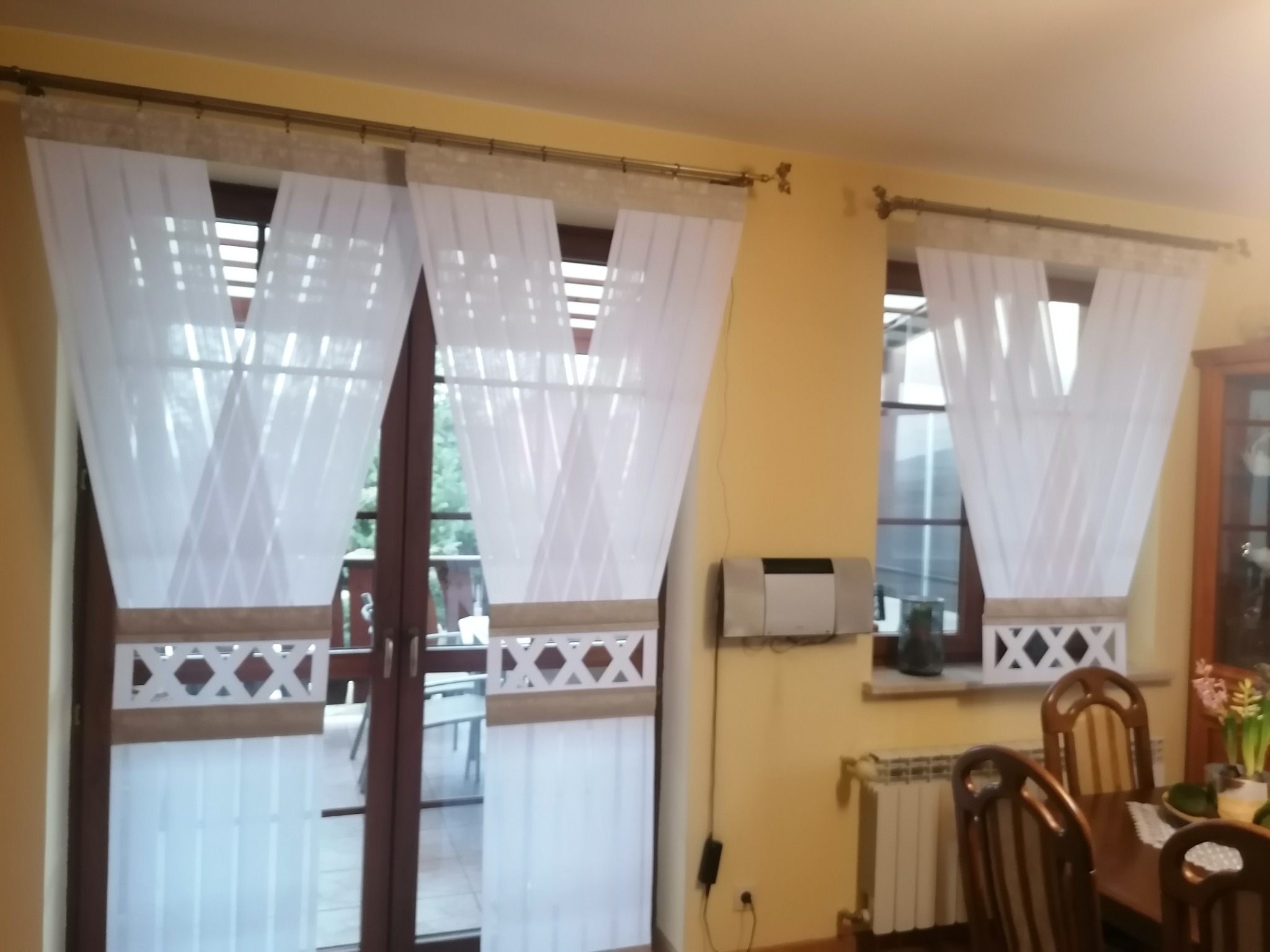 Białe, częściowo transparentne zasłony z dekoracyjnym pasem w geometryczny wzór, zawieszone na karniszu w salonie z żółtymi ścianami.