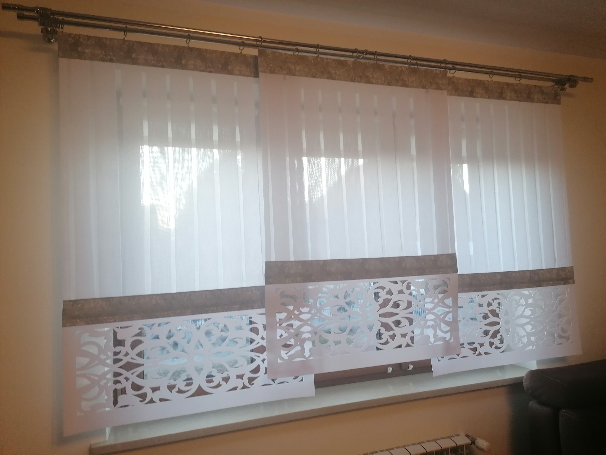 Białe, pionowe żaluzje z dekoracyjnym, ażurowym wykończeniem na dole, zawieszone na metalowym karniszu z ozdobnym pasem materiału u góry.