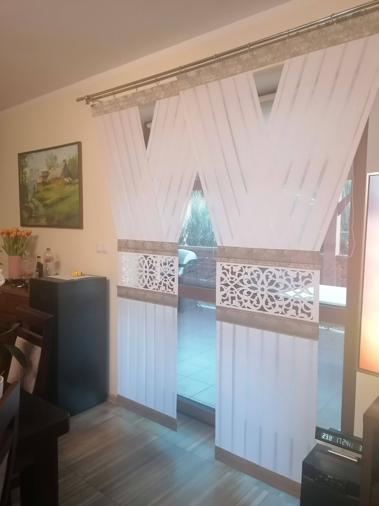 Eleganckie, białe zasłony panelowe z dekoracyjnym, ażurowym wzorem, zawieszone na karniszu w jasnym wnętrzu, z widokiem na zewnątrz.