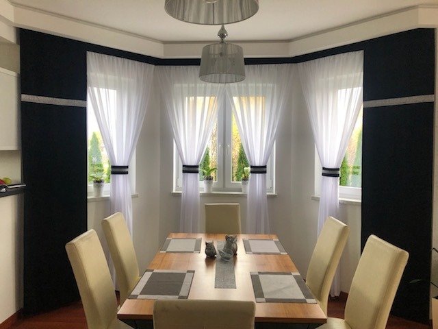 Jadalnia z drewnianym stołem i krzesłami, w oknach białe firanki z czarnymi akcentami i czarne zasłony z srebrnym paskiem, widok na ogród