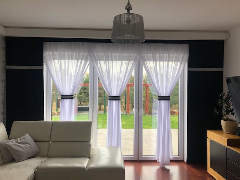 Salon z białymi, zwiewnymi zasłonami upiętymi czarnymi pasami, widok na ogród przez przeszklone drzwi balkonowe, skórzana sofa w rogu.